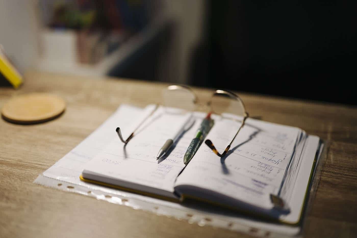 An open notebook with two pens and a pair of glasses on a wooden desk